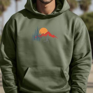 May include: A green hooded sweatshirt with a graphic design of a mountain range, a sun, and trees. The graphic is in shades of red, orange, blue, and white.