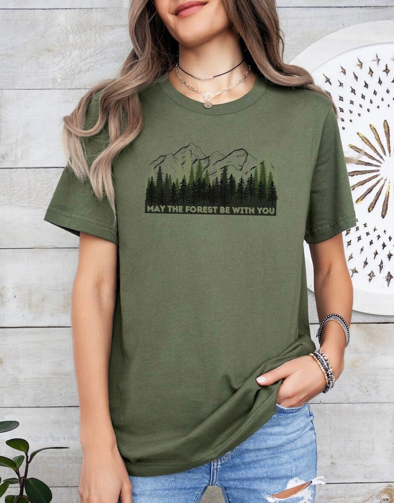 May include: A green t-shirt with a graphic of a mountain range and trees. The text "May the forest be with you" is printed below the graphic.