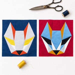 May include: Two square fabric quilt blocks featuring geometric animal face designs. One block has a dark blue background with a white, orange, and yellow fox-like face. The other has a red background with a light blue, yellow, and black face.