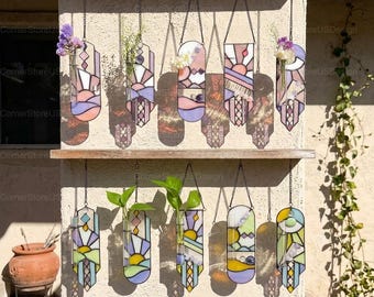 May include: Ten stained glass wall hangings with integrated glass test tube vases. Each piece features geometric patterns in pastel shades of pink, purple, blue, and yellow, suspended by metal chains from two wooden shelves.
