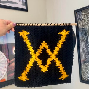 May include: A black and yellow crocheted wall hanging with a geometric design. The hanging is attached to a wooden dowel rod.