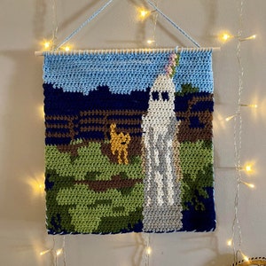 Op de afbeelding: Een gehaakte wandhanger met een geest met een bloem op zijn hoofd, een klein geel wezen en een blauw, groen en bruin landschap.