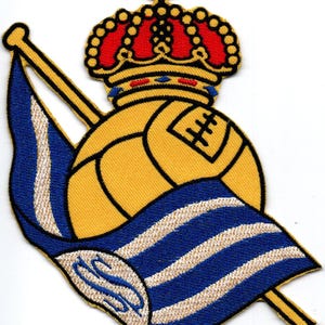 Pode incluir: Um remendo bordado que apresenta uma bola de futebol amarela com uma bandeira listrada azul e branca cobrindo-a. Uma coroa com detalhes vermelhos e azuis se assenta sobre a bola.