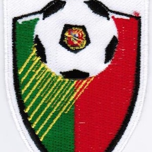 May include: Embroidered patch with the word "LIGA" at the top. The patch features a soccer ball over a shield divided into green and red sections. The soccer ball has a small emblem in the center.