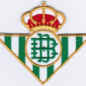 Puede incluir: Un parche triangular verde y blanco con una corona bordada en oro y una letra 'B' bordada en oro dentro de un círculo. El parche es un logotipo de equipo.