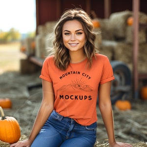 May include: A woman wearing a burnt orange t-shirt with a black graphic design that says "Mountain City Mockups" and a mountain range with a sun rising behind it. She is sitting on a bale of hay with pumpkins in the background.