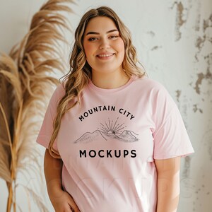 May include: A light pink t-shirt with the text "MOUNTAIN CITY MOCKUPS" in black. The design features a mountain range with a rising sun. The shirt is short-sleeved and has a round neckline.