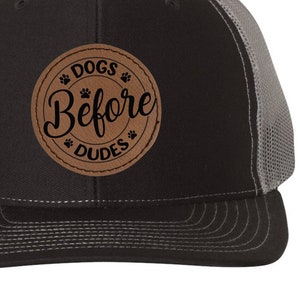 May include: A black and gray trucker hat with a brown leather patch that says "Dogs Before Dudes" in black lettering.