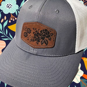 以下が含まれることがあります： グレーと白のトラッカーハットに、茶色の革のパッチが付けられており、花柄のデザインが施されています。ハットはメッシュバックで、カーブしたつばが付いています。