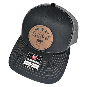 Może przedstawiać: Czarna i szara czapka trucker z brązową skórzaną naszywką z napisem "Body By Brisket" z sylwetką krowy i imieniem "Nick". Czapka jest regulowana i ma logo Richardson 112.