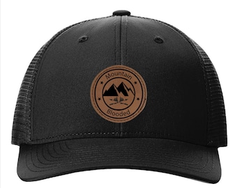 May include: Black trucker hat with a brown leather patch that says "Mountain Blooded" with a mountain silhouette and crossed axes.