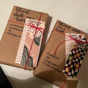 May include: Two brown paper-wrapped packages with red string tied around them. Each package has a sticker with the text "blind date with a book" and a bookmark with details about the book inside. One bookmark has a black and white checkered pattern with smiley faces and the other has a pink and black abstract design.