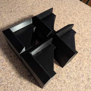 May include: A set of four black, geometric, 3D-printed objects. Each object has a trapezoidal shape with a flat top and angled sides. The objects are arranged together, creating a unique, abstract form. The surface appears smooth with visible layer lines.