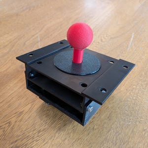 May include: Black arcade joystick with a red ball top. The joystick has a square base with mounting holes and a circular plate around the base of the stick. The joystick is designed for use in arcade games.