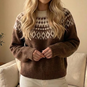 May include: A brown sweater with a white and brown geometric pattern across the chest and shoulders. The sweater has a crew neck and long sleeves. The person is wearing white pants. The background is a neutral color.