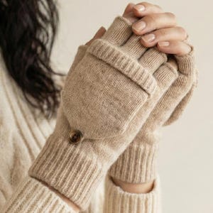 May include: Beige convertible mittens with a button closure. The mittens have a ribbed cuff and a flap that can be flipped back to expose the fingers. The mittens are made of a soft, knitted material.