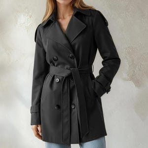 Può includere: Un trench nero con design a doppio petto, cintura e colletto a revers. Il cappotto ha maniche lunghe e viene indossato con jeans azzurri. Lo sfondo è un muro neutro e strutturato.