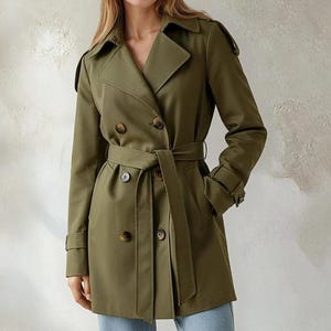May include: An olive green trench coat with a double-breasted design, a matching belt, and button details. The coat has long sleeves and a classic collar. The model is wearing light blue jeans.