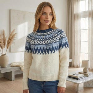 May include: A cream-coloured jumper with a round neck and long sleeves. The upper section features a blue and white geometric pattern. The model is wearing blue jeans. The jumper is made of a soft, warm material.