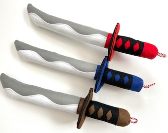 May include: Three gray felt toy swords with different colored handles. The top sword has a red handle with black diamond shapes. The middle sword has a blue handle with black diamond shapes. The bottom sword has a brown handle with black diamond shapes.