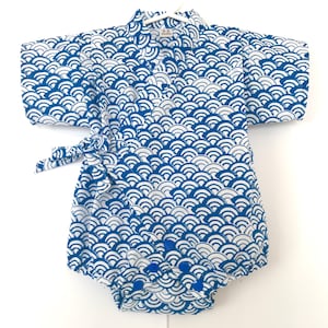 May include: A white and blue baby onesie with a Japanese wave pattern. The onesie has short sleeves and a wrap-around tie closure.