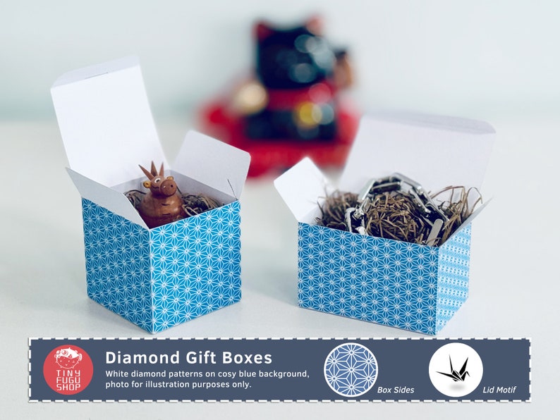 以下が含まれることがあります： 白い蓋と青いダイヤモンド模様の、開いた正方形のギフトボックスが2つ。1つの箱には小さな茶色の置物、もう1つには金属製のオブジェクトが入っています。画像には「Diamond Gift Boxes」と書かれています。