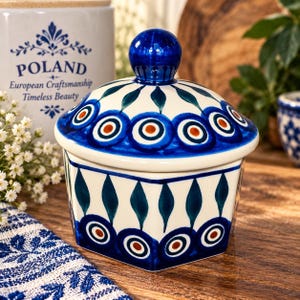 May include: A hexagonal ceramic jar with a matching lid featuring a blue, teal, and orange peacock-eye pattern on a cream background. The lid has a round blue knob handle. The jar displays a repeating geometric motif of stylized leaves and circles.