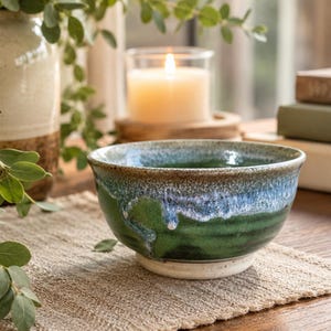May include: A ceramic bowl featuring a reactive glaze with a deep green base, transitioning into a mottled blue and white rim. The bowl has a rounded shape with a flared opening and a solid, unglazed off-white circular base at the bottom.
