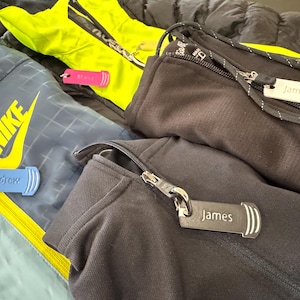 Puede incluir: Primer plano de varias chaquetas con cremalleras y etiquetas con nombres. Una chaqueta es azul marino con un logotipo Nike amarillo y una etiqueta azul que dice "Andrew". Otra chaqueta es negra con una etiqueta que dice "James".