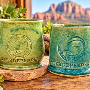 May include: Two ceramic mugs with a glossy finish in shades of light green and teal. Each mug features a circular embossed emblem with a swirling design and the word INTERDEPENDENCE moulded into the base. The mugs have sturdy handles.