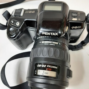 May include: A black Pentax PZ-10 camera with a Pentax 28-80mm zoom lens. The camera has a digital display and a black strap.