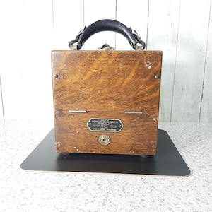 Può includere: Una valigetta portatile vintage in legno Weston Electrical Instrument Co. Modello 689. La valigetta ha un manico in pelle nera, ferramenta in metallo e una piccola targhetta metallica con testo. Il legno ha una venatura naturale e un aspetto usurato.
