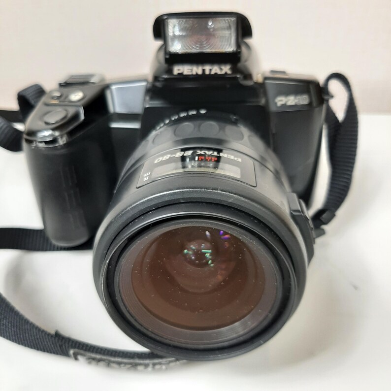 May include: A black Pentax PZ-1S 35mm film camera with a black Pentax 28-80mm lens attached. The camera has a built-in flash and a black strap.