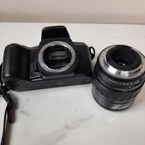 May include: A black 35mm film camera with a black lens attached. The camera has a silver lens ring and a silver ring around the lens mount. The lens has a silver ring with numbers and markings.