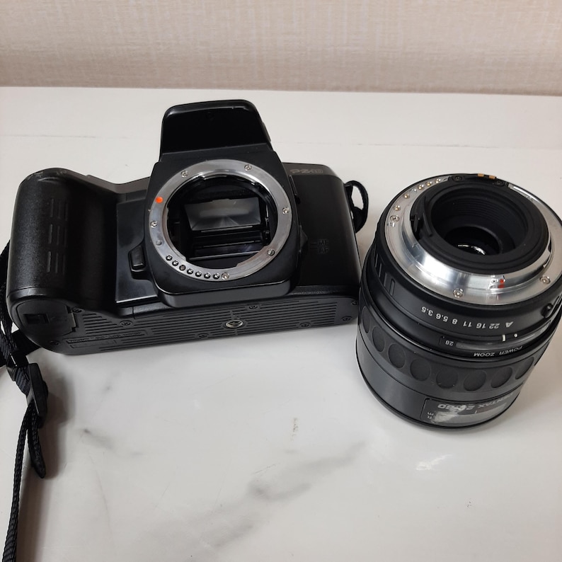 May include: A black 35mm film camera with a black lens attached. The camera has a silver lens ring and a silver ring around the lens mount. The lens has a silver ring with numbers and markings.