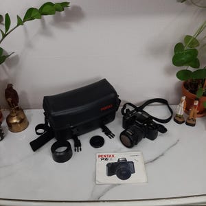 May include: A black Pentax Pz-10 camera with a lens attached, a black camera bag, a lens cap, and a manual. The camera has a black strap attached.