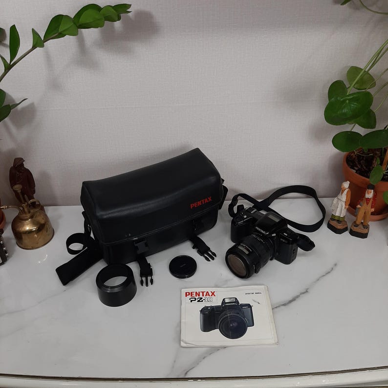 May include: A black Pentax Pz-10 camera with a lens attached, a black camera bag, a lens cap, and a manual. The camera has a black strap attached.