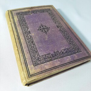 May include: An old book with a purple cover and a decorative black border. The cover has a cross design in the centre. The spine and edges are a light tan colour, showing signs of wear and age.