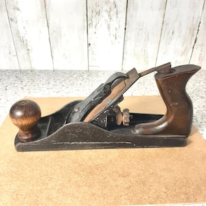 Könnte beinhalten: Ein alter Handhobel mit dunklem Holzgriff und Knauf, einem schwarzen Metallkörper und einer kupferfarbenen Klinge. Das Werkzeug ist für die Holzbearbeitung konzipiert und verfügt über einstellbare Einstellungen.