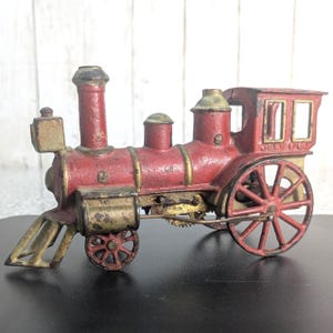 May include: A vintage cast iron toy train engine painted in red with gold-toned accents. The locomotive features a cowcatcher, a smokestack, a cab with open windows, and spoked wheels. The text "JUNE 1 76" is embossed on the side of the cab.