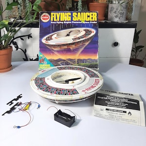 Op de afbeelding: Vintage Cox Flying Saucer speelgoed met originele doos en accessoires. De doos heeft een kleurrijke illustratie van de vliegende schotel. Het speelgoed is wit, rood en blauw met een zwarte propeller. Inclusief instructies en batterijhouder.