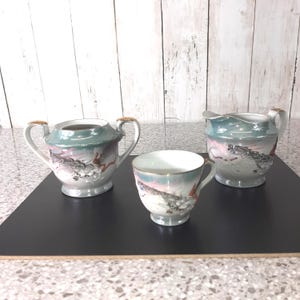 Op de afbeelding: Een driedelige porseleinen theeset met een drakenmotief. De set bevat een suikerpot, een melkkan en een theekopje. De stukken zijn wit met een turquoise en roze design en gouden accenten.