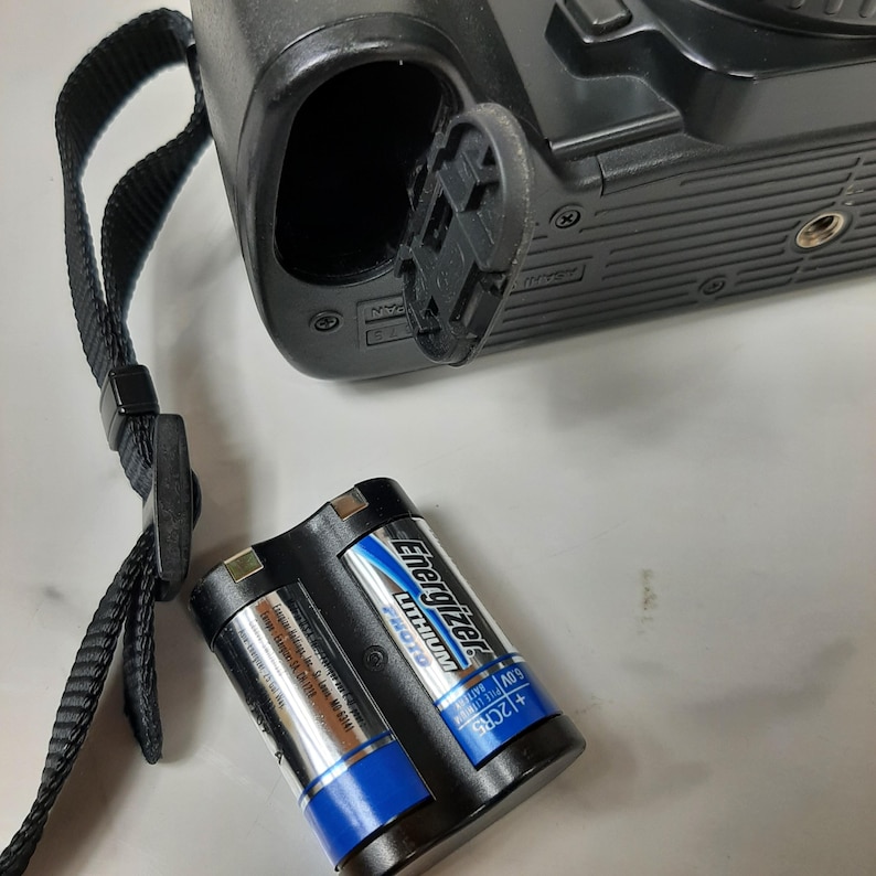 May include: A black camera with a battery compartment open, revealing two AA batteries labeled "Energizer MAX" and "1.5V Alkaline".