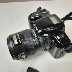 May include: A black Pentax P2-10 film camera with a Pentax 28-80mm zoom lens attached. The camera has a digital display on the top and a silver button on the side. The lens has a silver ring with the text "POWER ZOOM" and numbers 50 and 80.