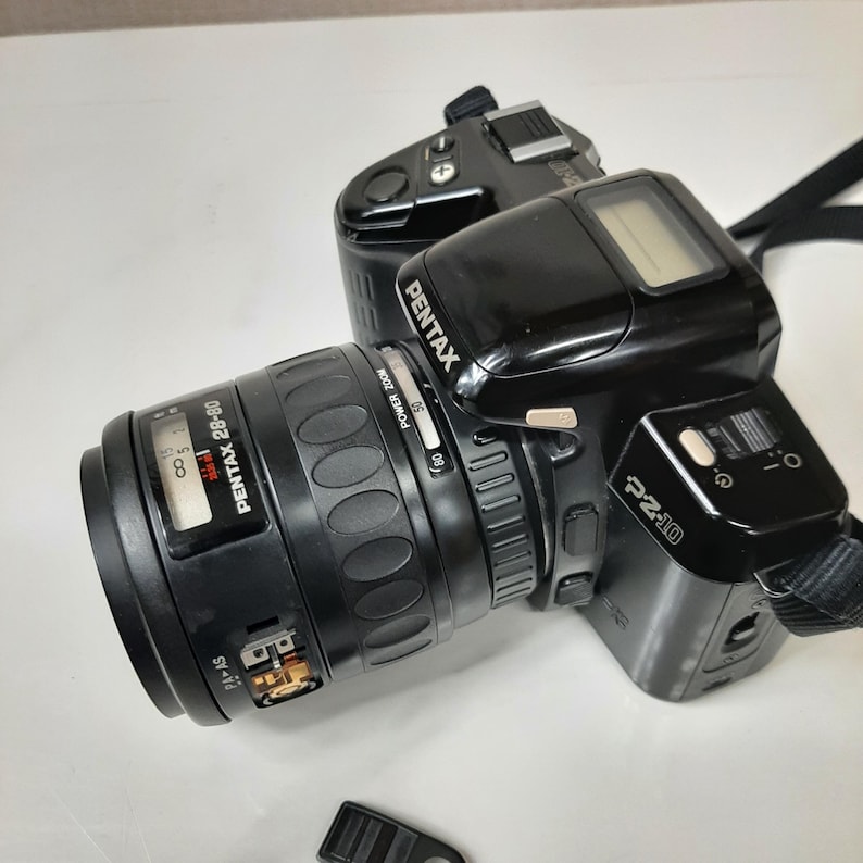 May include: A black Pentax P2-10 film camera with a Pentax 28-80mm zoom lens attached. The camera has a digital display on the top and a silver button on the side. The lens has a silver ring with the text "POWER ZOOM" and numbers 50 and 80.