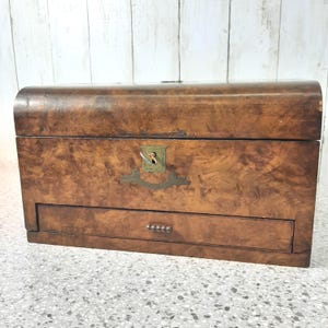 Puede incluir: Un cofre de madera vintage con una parte superior curvada y un cajón pequeño. El cofre está hecho de madera oscura estampada con herrajes de latón, que incluyen una cerradura y un tirador de cajón. El cofre probablemente se usa para almacenamiento.