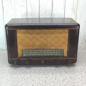 May include: A vintage tabletop radio with a dark brown, polished bakelite casing and a textured, gold-coloured fabric speaker grille. The front features a horizontal glass tuning dial with printed frequency scales and four circular control knobs.