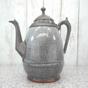 May include: A grey and white mottled enamel coffee pot featuring a metallic grey lid, handle, and spout. The body has a rounded shape with a copper-coloured base. The surface displays a speckled pattern throughout, and the lid is topped with a small knob.