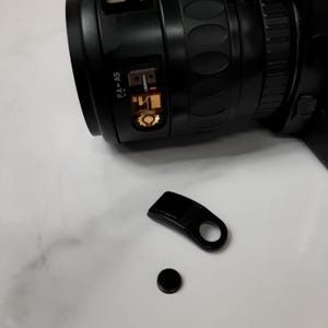 May include: A black camera lens with a gold-colored ring and the text "PA-AS" on the side. The lens is detached from the camera and is lying on a white surface. There is a black plastic lens cap and a black plastic lens hood next to the lens.