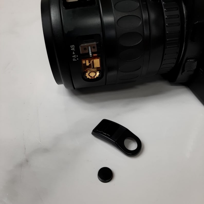 May include: A black camera lens with a gold-colored ring and the text "PA-AS" on the side. The lens is detached from the camera and is lying on a white surface. There is a black plastic lens cap and a black plastic lens hood next to the lens.